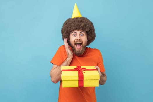 Portrait of excited man with Afro hairstyle wearing orange T-shirt and party cone holding his birthday present, being surprised, yelling. Indoor studio shot isolated on blue background.