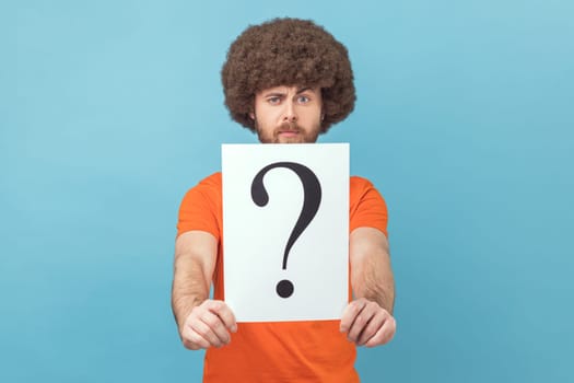 Portrait of man with Afro hairstyle wearing orange T-shirt holding question mark, finding smart solution, asking for advice, looking at camera. Indoor studio shot isolated on blue background.