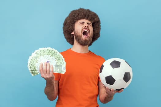 Excited man with Afro hairstyle wearing orange T-shirt holding soccer ball and hundred euro bills, screaming with happiness, betting and winning. Indoor studio shot isolated on blue background.