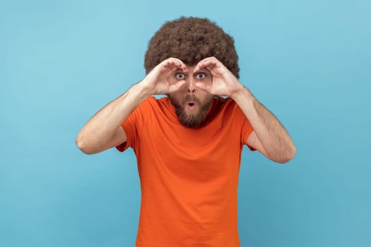 Portrait of man with Afro hairstyle wearing orange T-shirt holding fists near eyes imagining binoculars and looking through holes, spying, having fun. Indoor studio shot isolated on blue background.