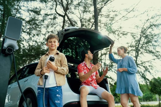 Little boy holding EV charger and point at camera with his family sitting on the trunk in background. Road trip travel with alternative energy charging station for eco-friendly car concept. Perpetual