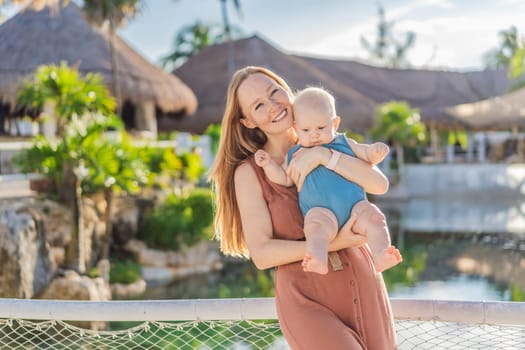 Amidst tropical palms and thatched roofs, a loving mom embraces her baby, sharing warmth and affection in a tranquil outdoor setting.