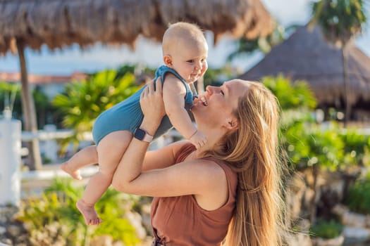 Amidst tropical palms and thatched roofs, a loving mom embraces her baby, sharing warmth and affection in a tranquil outdoor setting.