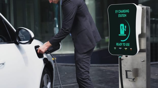 Businessman recharge his electric car from charging station at city center or public car park. Eco friendly rechargeable car using alternative clean energy in urban city lifestyle.Peruse