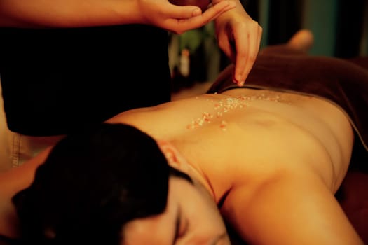 Man customer having exfoliation treatment in luxury spa salon with warmth candle light ambient. Salt scrub beauty treatment in Health spa body scrub. Quiescent