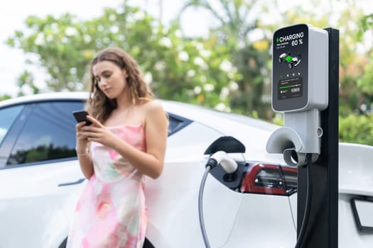 Focused charging station recharging electric vehicle on blurred background of modern woman using smartphone. EV technology utilization for tracking energy usage to optimize battery charging.Synchronos