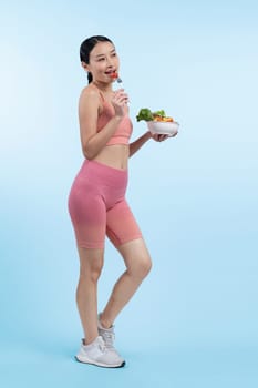 Young sporty Asian woman in sportswear holding salad bowl fill with vibrant of fruit and vegetable. Natural youthful and fit body lifestyle with balance nutrition on isolated background. Vigorous