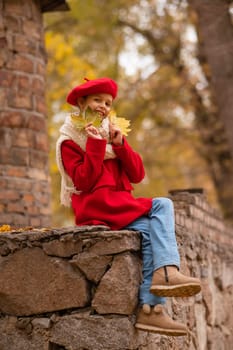 Smiling caucasian girl in a red coat and beret sits on a brick wall on a walk in autumn