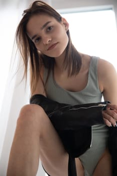 a woman in a gray bodysuit and a black jacket stands by the window