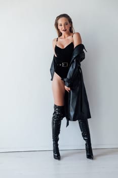 a woman in a leather cloak and black clothes stands against a white wall