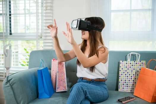 Happy young woman customer wearing 3d VR glasses virtual reality headset with shopping bags around she touching and pointing on air to online shopping in living room at home, digital cyberspace