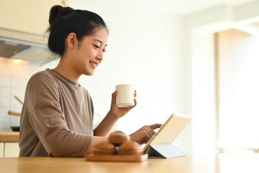 Attractive young woman drinking coffee and using digital tablet at table in kitchen.