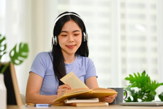 Charming young woman in headphone reading book, enjoying leisure weekend time at home.