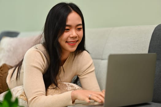 Happy young woman in warm sweater using laptop while lying on couch at home.