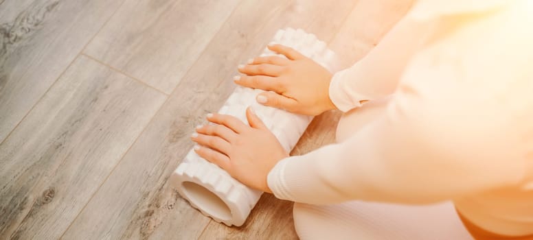 Adult athletic woman, in white bodysuit, performing fascia exercises on the floor - caucasian woman using a massage foam roller - a tool to relieve tension in the back and relieve muscle pain - the concept of physiotherapy and stretching training.