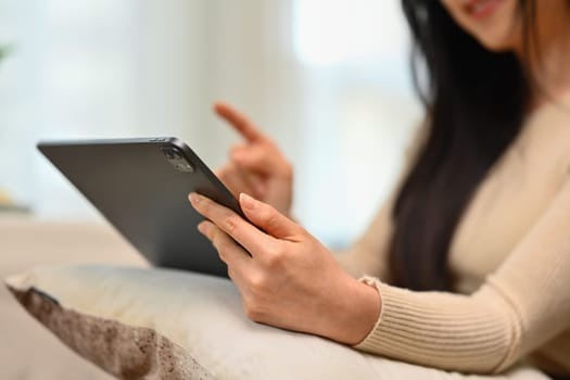 Cropped shot of young woman in warm sweater using digital tablet on couch at home.