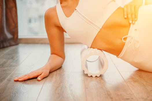 Adult athletic woman, in white bodysuit, performing fascia exercises on the floor - caucasian woman using a massage foam roller - a tool to relieve tension in the back and relieve muscle pain - the concept of physiotherapy and stretching training.