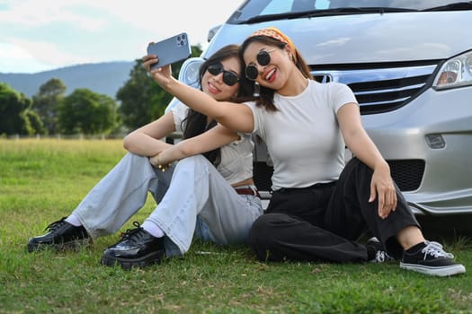 Cheerful female friends taking a break from driving siting in front of car on meadow and taking selfies with smartphone.