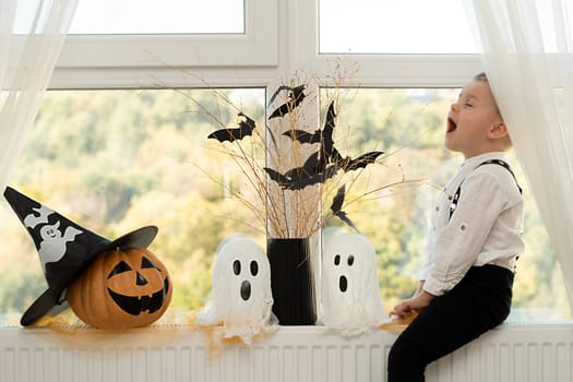 Halloween. Concept. A cute and cheerful boy sits near the window against the background of white ghosts and a vase with dry branches and black paper bats.Close-up.