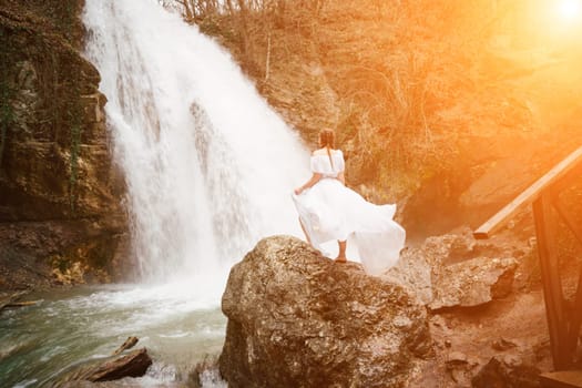 Happy woman in a white dress stands on a stone with a waterfall behind.