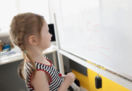 Portrait of toddler writing solution of sums on white board at school, educational process. School kid think while doing mathematics problem. Learn concept