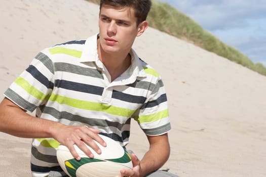 Teenage Boy Sitting On Beach Holding Rugby Ball