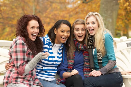 Group Of Four Teenage Girls Sitting On Bench In Autumn Park