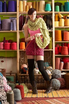 Young Woman Knitting Scarf Standing In Front Of Yarn Display