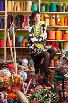Young Woman Sitting On Stool Holding Knitting Needles In Front Of Yarn Display