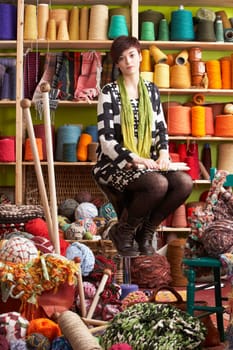 Young Woman Sitting On Stool Holding Knitting Needles In Front Of Yarn Display