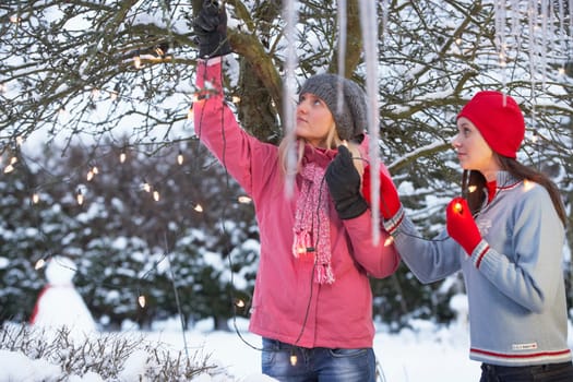 Two Teenage Girls Hanging Fairy Lights In Tree With Icicles In Foreground