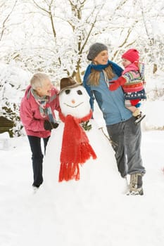 Young Girl With Grandmother And Mother Building Snowman In Garden