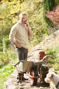Man Outdoors On Mobile Phone With Dog Whilst On Break From Gardening