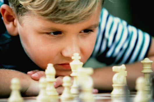 Cropped close-up of a young boy resting his head on his hands as he studies a chess board. Horizontal shot.