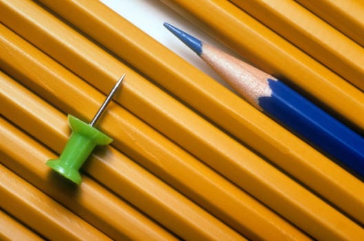 Standard pencils and a sharpened blue pencil are lined up next to each other. There is a green pushpin on top of the pencils. Horizontal shot.