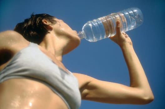 Low angle view of a woman in exercise gear, drinking water from a water bottle against a blue sky. Horizontal shot.
