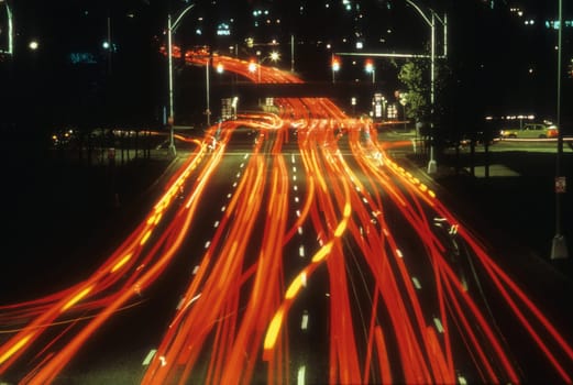 Tail lights of cars are seen at night as red lines as they pass along a street through an intersection. Horizontal shot.