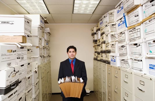 A young businessman is standing in a file room holding files. Cabinets and storage boxes line the sides. Horizontal shot.