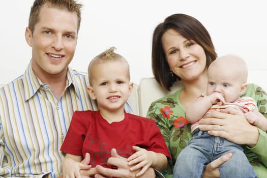 Family composed of a mother, father and two small children. They smile towards the camera while sitting on a sofa. Horizontal shot.