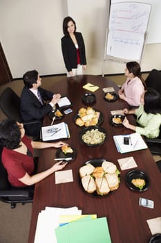 A group of business executives attend a meeting with food on the conference table. A woman stands at the head next to a dry erase board. Vertical shot.