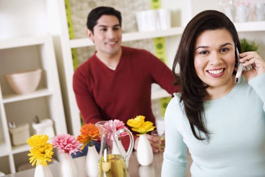 A young woman is talking on a cell phone while a young man stands behind a kitchen counter in the background. Horizontal shot.
