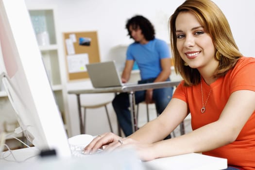 Young woman smiles towards the camera while at her computer. A man in the background works on his laptop. Horizontal shot.