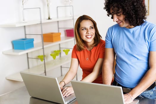 A young man and woman are smiling at one another while working side by side on laptops. Horizontal shot.