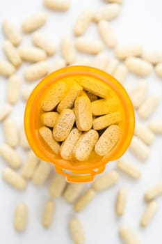 Overhead view of a yellow pill bottle full of tablets. Tablets are also scattered around on a white surface. Vertical shot.