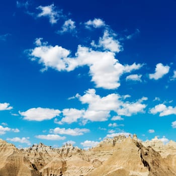 Mountain terrain in Badlands National Park, South Dakota, beneath blue sky and clouds. Square format.