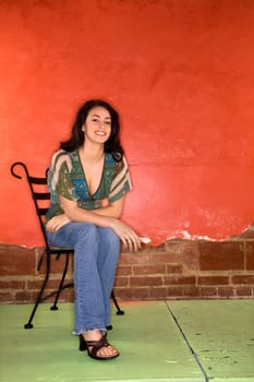 Attractive young adult woman sitting on a chair in front of a colorful wall. Vertical shot.