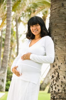 Portrait of a pregnant woman leaning against a tree and holding her belly. Vertical shot.