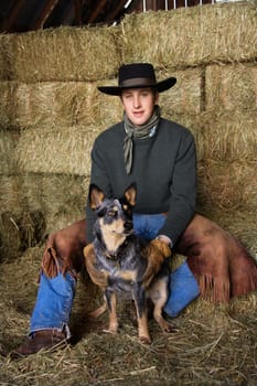 Attractive young man wearing a black cowboy hat, sitting atop a hay bale. He is smiling and holding an Australian Shepherd. Vertical shot.
