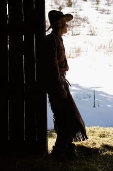 Silhouette of a cowboy leaning in a barn doorway, looking outside. Vertical shot.