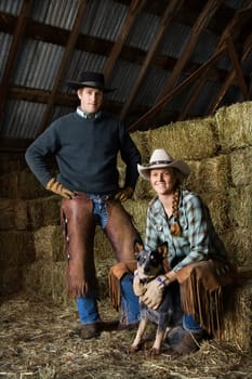 Attractive young man and woman with an Australian Shepherd in a barn. Bales of hay are in the background. Vertical shot.